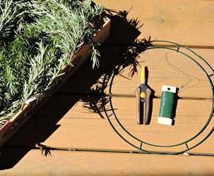 box of rosemary sprigs-branch tips laying next to a wire wreath frame, scissors and paddle wire. 