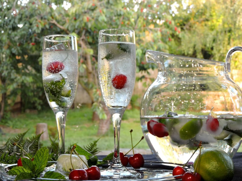 cherry ice cubes floating in sparkling mineral water with mint and lime ice cubes and cherry tree in background